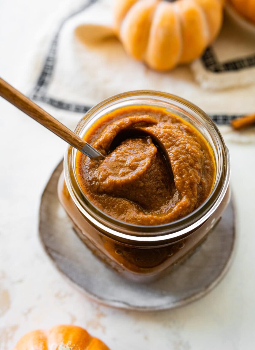 A glass jar full of pumpkin butter with a spoon inside.