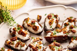 A large plate of multiple dates that are stuffed with goat cheese and topped with chopped pecans and fresh thyme.