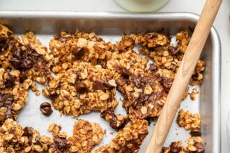 Clusters of chocolate chip cookie granola on a silver baking sheet served with a wooden spoon.