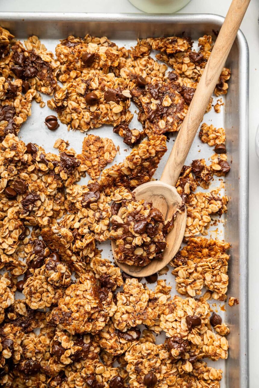 Clusters of chocolate chip cookie granola on a silver baking sheet served with a wooden spoon.