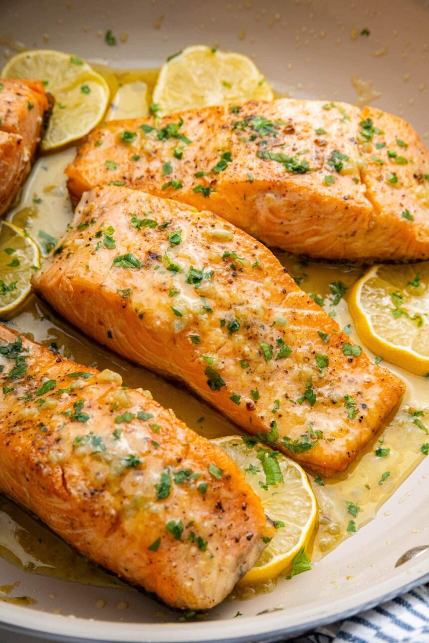 Four salmon filets in a pan with garlic butter and lemon wedges. The filets are garnished with fresh herbs.