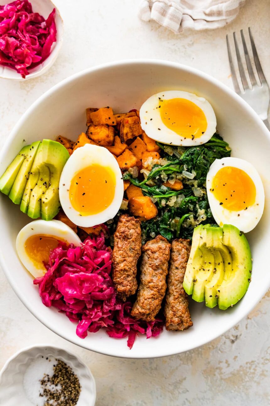 A breakfast bowl with 2 soft boiled eggs, 3 turkey sausage, sliced avocado, sauerkraut, roasted sweet potatoes and sautéed kale all in a white bowl.