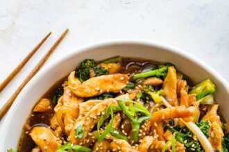 Chicken and broccoli stir fry in a white bowl, garnished with sesame seeds and green onions, with wooden chopsticks beside it.