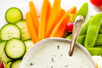 Cottage cheese ranch dip in a white bowl garnished with fresh dill. The bowl is on a plate with an assortment of fresh vegetables.