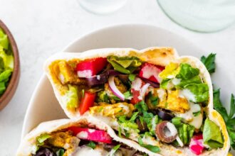 Three halves of chicken shawarma pita pockets stacked in a bowl, filled with veggies, olives, pickled turnips, and topped with yogurt sauce and fresh parsley.
