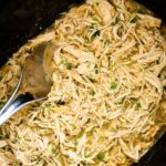 Shredded salsa verde chicken in a slow cooker with a silver serving spoon. Fresh cilantro is on top of the chicken.
