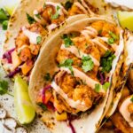 Three shrimp tacos in flour tortillas folded up on a serving platter. The tacos are topped with a mango cabbage slaw and a drizzle of spicy crema.